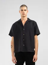 Camisa negra de manga corta con cuello camp y calce relajado. Confeccionada en una mezcla de lino y algodón, terminada con botones de madera y parche característico de Polonio. Hecha en Italia.