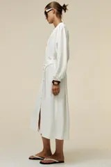 Vestido midi blanco de crepe con cuello redondo y escote pico, manga larga con puños y cinturón de la misma tela con hebilla metálica. Cierre frontal con botones metálicos.