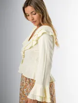 Blusa color crudo en lino rayón con volados, de mangas largas acampanadas y cierre frontal con botones.
