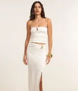 Conjunto de dos piezas color blanco, compuesto por un top tipo corset strapless con escote en V y una pollera midi ajustada con fruncido lateral y abertura alta, asegurada con un adorno metálico.