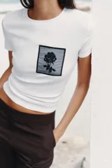 Camiseta blanca de algodón con cuello redondo y mangas cortas. Presenta un bolsillo de tul con una flor bordada y abalorios en el delantero.