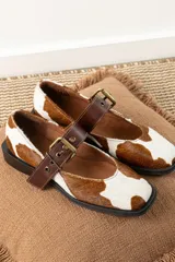 Zapatos Mary Jane con estampado de vaca en blanco y marrón, correa de cuero marrón con hebilla dorada y suela plana de goma negra.