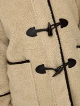 Abrigo de tela tipo teddy color beige, con capucha, cierre con botones tipo duffle coat y bolsillos frontales. Detalles en cuero sintético negro en los bordes y en los botones.
