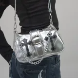 Cartera de hombro plateada con acabado metálico, diseño tipo baguette. Presenta dos apliques de cruces negras con pespuntes blancos en el frente y una hebilla decorativa central. Incluye correa ajustable y cadena metálica plateada.