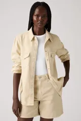 Camisa de manga larga color beige claro, estilo western, con cuello y botones a presión frontales. Presenta dos bolsillos de parche con solapa en el pecho y un pequeño tag rojo en el bolsillo derecho.