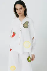 Camisa blanca de lino con corte raglán, detalles desflecados y apliques de frutas tejidas a crochet. Abertura trasera con botones.
