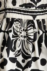 Vestido corto sin mangas de algodón color crudo con estampado de flores, hojas y lunares en negro. Tiene escote cruzado en V, cintura elastizada y falda con vuelo.