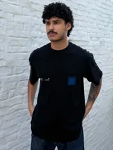 Remera negra de corte oversize con cuello redondo y manga corta. Presenta un pequeño estampado blanco con texto y un cuadrado difuminado azul en el pecho.