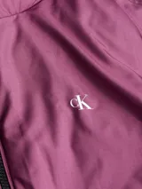 Campera reversible de poliéster impermeable color borgoña con logo Calvin Klein Jeans estampado en blanco. Un lado es liso y el otro tiene un diseño con monogramas.