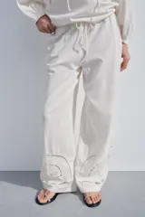 Pantalón de tiro medio color crudo, confeccionado en mezcla de algodón y lino, con cintura elástica ajustable con cordones, bolsillos laterales ocultos y detalle de bordados perforados a tono en el ruedo.