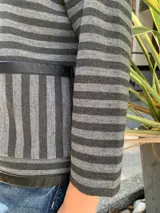 Chaqueta tejida en lana merino a rayas horizontales grises y negras, con ribete de cuero negro en cuello, cierre y bolsillos.