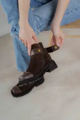 Bota corta de cuero gamuzado color marrón chocolate, con punta cuadrada y suela track. Presenta una correa con hebilla desmontable y cierre lateral con cremallera.