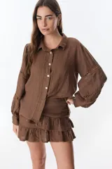 Camisa color marrón de lino, con cuello camisero, cierre frontal con botones, mangas largas abullonadas con detalle de encaje y corte holgado.
