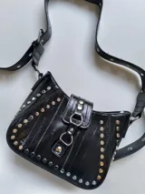 Cartera negra de charol con forma de media luna y tachas plateadas. Tiene correa de hombro ajustable y cierre con hebilla.