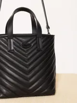 Cartera tote pequeña de cuero sintético negro con diseño acolchado en forma de espiga. Tiene doble asa corta superior, correa larga ajustable y desmontable, y logo metálico frontal con forma de labios.