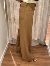 Pantalón ancho color marrón claro, de corte holgado, con pinzas debajo de la rodilla, bordado de monograma en el bolsillo delantero y pespuntes a contratono.
