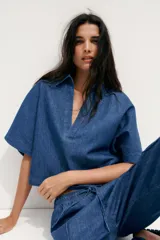 Camisa corta de denim azul, con cuello solapa, escote en pico y mangas cortas.