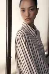 Camisa de crepé color crema con rayas verticales marrones, corte oversized, cuello clásico, cierre frontal con botones ocultos, mangas largas con puños abotonados, pliegue de caja invertido en la espalda y bajo recto.