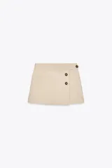 Falda pantalón color beige de tiro medio con diseño cruzado y cierre con botones. Cuenta con bolsillos falsos en la parte delantera y trasera, y cierre lateral con cremallera oculta.