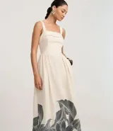 Vestido midi de corte evasé, color crema, con escote cuadrado y breteles anchos. Presenta un estampado grande de hojas y follaje en tonos oscuros en el ruedo inferior.