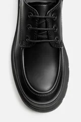 Zapato de vestir tipo blucher color negro, con detalle de bordón, tirador trasero y suela con volumen irregular.