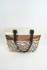 Bolso tipo tote con estampado animal print de cebra en tonos marrones y blancos, con una franja horizontal color bronce y asas de mano marrones.
