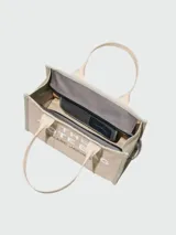Bolso tipo tote color beige con bordes grises y estampado frontal con el texto "THE TOTE BAG MARC JACOBS" en blanco.