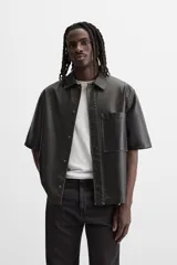 Sobrecamisa negra de manga corta confeccionada en tejido efecto piel, con cuello solapa, bolsillo frontal de parche y cierre frontal con botonadura oculta.