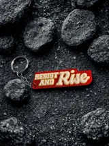 Llavero rectangular con cadena y aro metálico. Diseño con la frase "Resist and Rise" en letras blancas con borde amarillo sobre fondo rojo.