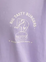 Camiseta lila de manga corta y cuello redondo, con estampado frontal de un elefante sobre una hamburguesa y el texto "Big Tasty Burgers".