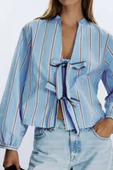Camisa celeste con rayas verticales marrones y blancas, cuello redondo con escote en pico y mangas largas abullonadas. Cierre frontal con lazos con pespuntes marcados.
