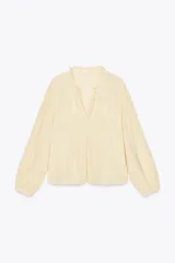 Blusa color crema de escote pico con volantes y manga larga. Detalle de jaretas y entredoses a tono. Cierre frontal con botones forrados a tono.