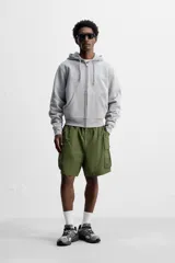 Bermuda color verde militar, relaxed fit, confeccionada en tejido técnico ligero. Cintura elástica ajustable con cordón interior, pliegues frontales, bolsillos delanteros y traseros con solapa, bolsillos cargo con solapa en perneras. Cierre frontal con cremallera y botón.