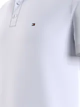 Chomba blanca de corte slim con cuello mao, tapeta de tres botones y logo de Tommy Hilfiger bordado en el pecho. El cuello presenta una banda con los colores característicos de la marca: azul marino, blanco y rojo.