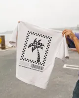 Remera blanca de manga corta con estampado en la espalda de una palmera dentro de un marco a cuadros y texto en la parte inferior.
