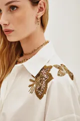 Camisa blanca de algodón con cuello clásico, mangas largas abullonadas con puños anchos y cierre frontal de botones. Presenta aplicaciones bordadas en dorado con detalles de animal print en los hombros.