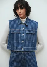Chaleco crop de jean azul con rayas verticales blancas, cuello clásico, cierre con botones y bolsillos frontales con solapa. El corte es recto y llega hasta la cintura.