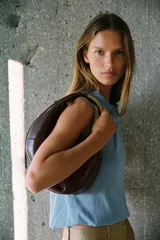 Bolso de mano ovalado color marrón oscuro, con asa de hombro metálica dorada y cierre superior con cremallera.