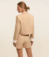 Blazer color beige con rayas verticales finas, cuello con solapa, doble botón, bolsillos delanteros y mangas largas con detalle contrastante en blanco.