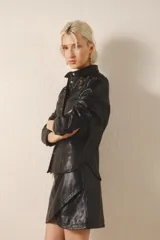 Camisa negra de cuero ovino con corte jeanero entallado, bolsillos tipo ojal y cierre frontal con broches a presión. Presenta parches bordados con canutillos y mostacillas en el frente.