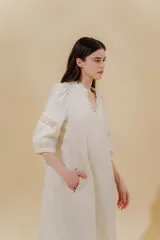 Vestido largo color beige, confeccionado en lino, con escote en V con volados y apliques de puntilla en las mangas. Posee bolsillos delanteros y tajos laterales.