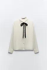 Camisa color crema de satén con cuello solapa, manga larga y lazo negro contrastante. Cierre frontal con botones.
