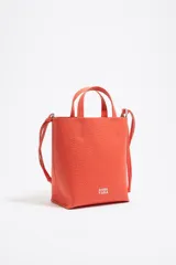 Bolso de mano mediano marca Bimba y Lola, color naranja, confeccionado en cuero con textura granulada. Tiene doble asa corta, correa bandolera ajustable y extraíble, logo estampado en el frente y herrajes plateados. El interior cuenta con etiqueta de tejido con el logo de la marca.