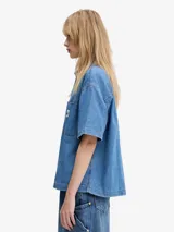 Camisa de denim azul claro de Carhartt WIP, corte relajado, manga corta, cuello camisero, cierre frontal con botones y bolsillo de parche en el pecho con logo de la marca.