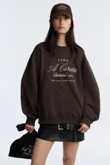 Sudadera negra de cuello redondo y manga larga, con bordado blanco en el frente que dice 'Zara A Coruña Established 1975 Rúa Juan Flórez 64-66' y un logo pequeño con el número 50 en la parte superior.