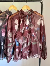 Camisa de seda italiana con estampado floral en tonos rosados, marrones y verdes. Presenta cuello alto con volados que caen en cascada sobre el frente y mangas largas abullonadas con puños ajustados.