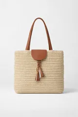 Bolso tipo tote bag de rafia tejida color beige, con asas y solapa superior de cuero marrón. Incluye un colgante decorativo con borlas de cuero.