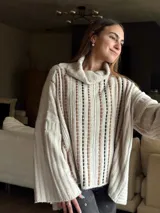 Sweater tejido color beige con cuello alto y mangas largas. Presenta aberturas verticales con detalles en marrón y negro.