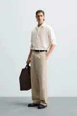 Pantalón de corte holgado (wide fit) en color beige, confeccionado en tejido con mezcla de lyocell. Presenta pliegues frontales en la cintura, bolsillos laterales y bolsillos traseros de doble vivo. Cuenta con cierre frontal de cremallera y botón.