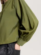 Camisa verde oliva de corte cropped, sin cuello, con cierre frontal de botones y mangas largas abullonadas con puños abotonados.